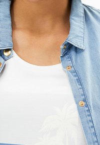 Chemise en jean bleu clair à boutons avec des détails dorés, portée sur un t-shirt blanc arborant un motif discret de palmier. Gros plan sur l'encolure.