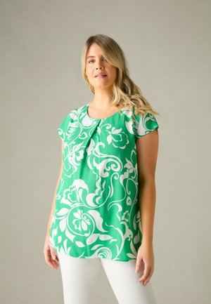 Blusa verde con motivo floreale e maniche corte, caratterizzata da un colletto rotondo e un dettaglio plisè sul davanti, indossata con pantaloni bianchi.