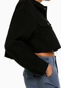 Svart kort jacka av bomull, med krage, knappstängning, två bröstfickor och slitna detaljer vid fållen.