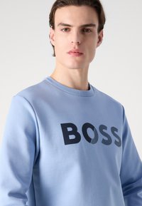 Light blue sweatshirt with a crew neck, featuring a bold black "BOSS" logo across the chest in a textured print. Smooth fabric finish.