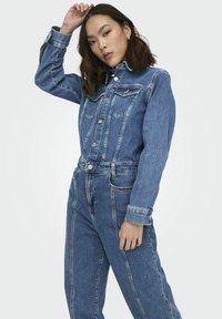 Jeune femme posant avec une main touchant sa tête, portant une veste en jean bleue et un pantalon en jean assorti sur un fond uni.