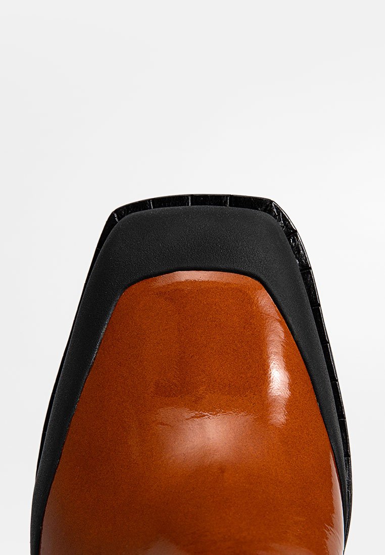 Brauner Lederschuh mit glänzender Oberfläche, quadratischer Zehenform, schwarzen strukturierten Akzenten und glattem schwarzen Absatz. Gravierte Details entlang der Kante.