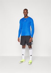 Kék hosszú ujjú sportpóló fekete rövidnadrággal párosítva, fehér zoknikkal fekete csíkokkal, és élénk sárga cipőkkel. Adidas logó látható.
