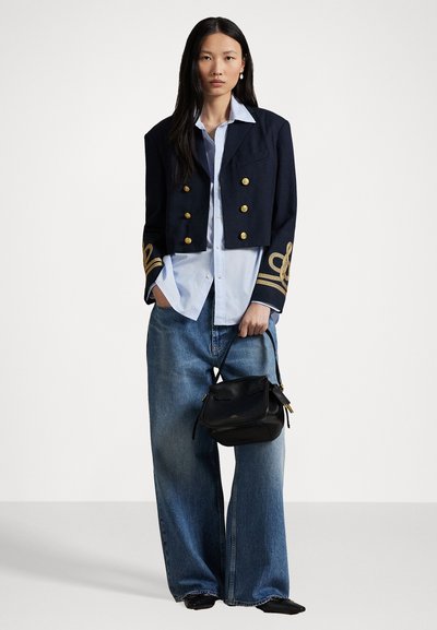 Navy cropped jacket with gold buttons and decorative shoulder accents over a light blue shirt and wide-leg denim jeans, holding a black handbag.