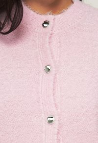 Απαλό ροζ αφράτο ζακέτα με στρογγυλή λαιμόκοψη, με πέντε λαμπερά ασημένια κουμπιά στον εμπρός μέρος και υφασμένη πλεκτή υφή.
