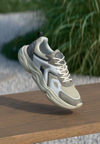 Sport Schuhe mit einem grauen und weißen Mesh-Obermaterial, gepolsterter Sohle, reflektierenden Akzenten und beigen Details, die über einer Holzoberfläche schwebt.