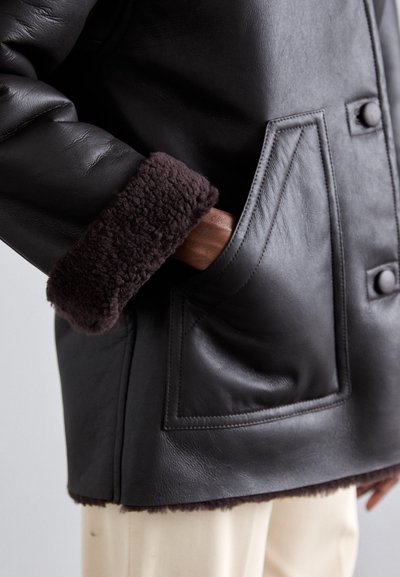 Crni kožni kaput s ovratnikom od smeđeg ovčjeg krzna i ukrasima na džepovima. Karakterizira ga glatka tekstura i strukturirani oblik.