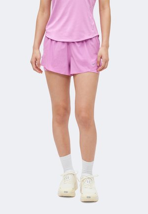Person iført lyse lilla træningsshorts og top, hvide sokker og cremefarvede sneakers, stående mod en ensfarvet baggrund.