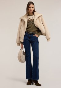 Chaqueta de felpa oversized en tono beige claro, suéter verde con estampado, jeans acampanados azules, botas de tacón verde oscuro y un pequeño bolso de pelo gris.