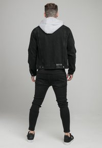 Man met nektatoe die een zwart denimjack met grijze capuchon draagt, zwarte jeans en zwarte sneakers, staand met de rug naar de camera tegen een effen achtergrond.