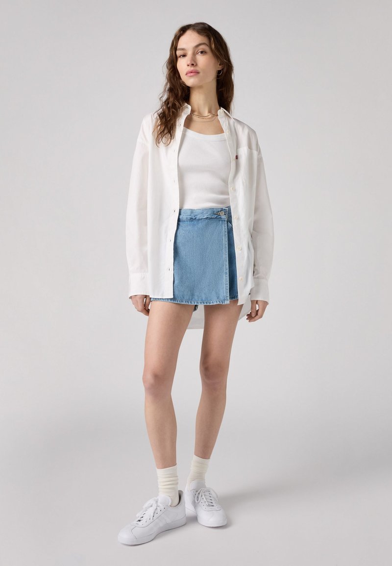 Weißes Button-Down-Hemd über einem weißen Tanktop, kombiniert mit einem blauen Jeans-Minirock und weißen Sneakers. Einfaches, lässiges Outfit.