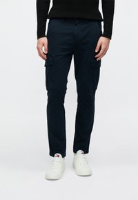 Μπλε σκούρο παντελόνι cargo με ίσια γραμμή, κατασκευασμένο από βαμβάκι. Διαθέτει πλαϊνές τσέπες και λεία υφή. Συνδυάζεται με λευκά αθλητικά παπούτσια.