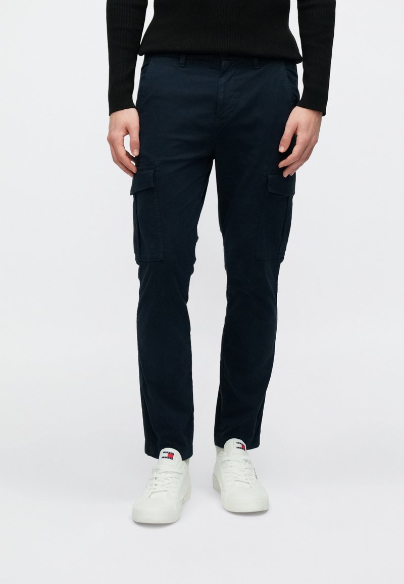 Μπλε σκούρο παντελόνι cargo με ίσια γραμμή, κατασκευασμένο από βαμβάκι. Διαθέτει πλαϊνές τσέπες και λεία υφή. Συνδυάζεται με λευκά αθλητικά παπούτσια.