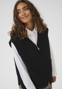 Crni flis prsluk s prednjim zatvaračem, dva velika džepa i dizajnom u obliku slova V, nosi se preko bijele košulje na dugmad. Jednostavna tekstura.