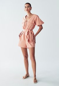 Persika värvi romper lühikeste puhvärvidega, sügava V-kaelusega, kahe ees oleva taskuga ja seotud vöökoht, valmistatud kergest kangast.