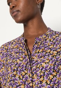 Camisa com padrão floral em roxo, laranja e amarelo sobre um fundo preto, apresentando um design de botão e um colarinho pregueado.