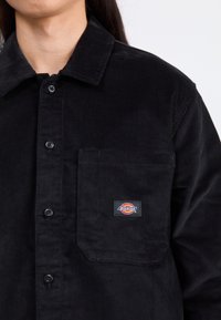 Camisa de pana negra con cierre de botones en la parte delantera, cuello puntiagudo y un único bolsillo en el pecho con una etiqueta de logo. Tejido texturizado.