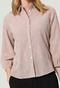Chemise légère de couleur rose poudré avec une texture côtelée verticale, fermeture boutonnée à l'avant et manches ballon trois-quarts. Présente un ourlet arrondi et un éclat subtil.