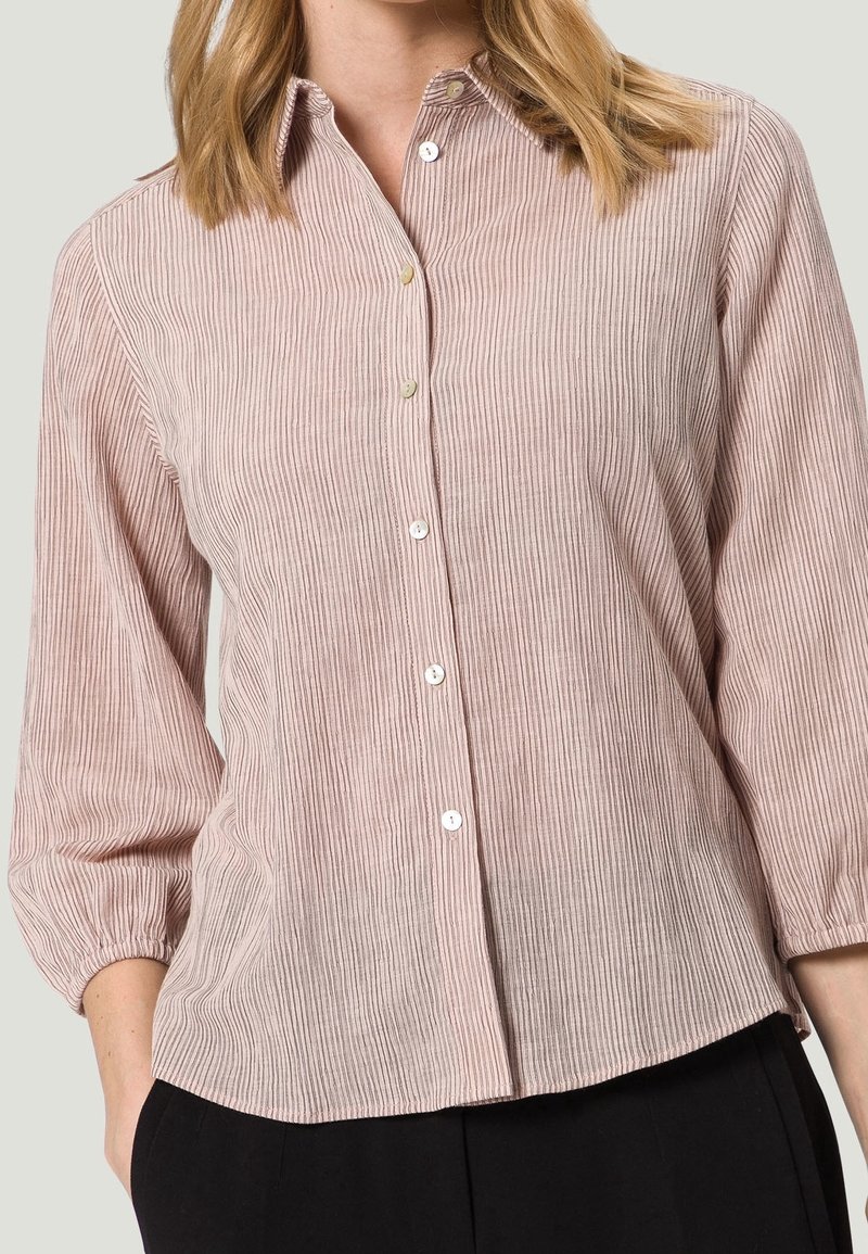 Chemise légère de couleur rose poudré avec une texture côtelée verticale, fermeture boutonnée à l'avant et manches ballon trois-quarts. Présente un ourlet arrondi et un éclat subtil.