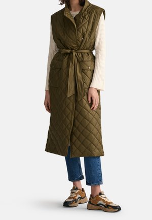 Vrouw draagt een mouwloze, olijfgroene gewatteerde lange bodywarmer met riem, een crèmekleurige gebreide trui, blauwe cropped jeans en tan zwarte sneakers.