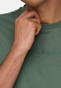Olive green crew-neck t-shirt with ribbed texture and embossed "ONLY & SONS" logo on the front. Model's hand is touching the neckline.