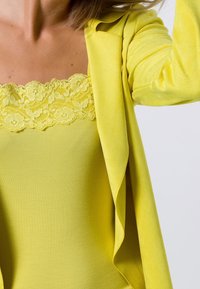 Żółta marynarka o miękkiej teksturze i luźnym kroju, zestawiona z pasującym żółtym topem z koronkowym wykończeniem przy dekolcie.