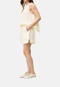 Helles gelbes zwei-teiliges Outfit mit einem ärmellosen Oberteil und passenden Shorts mit seitlichen Schlitzen. Die Schuhe sind hellbeige mit gebundenen Schnürsenkeln.