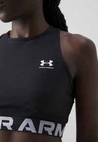 Černá sportovní podprsenka s vysokým výstřihem, s bílým logem "Under Armour" a kontrastním pásem s výrazným písmem na spodní části.