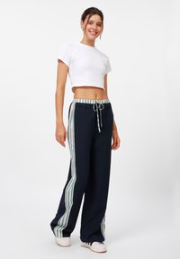 Marineblaue weit geschnittene Hose mit seitlichen Streifen in Grün und Weiß. Elastischer Bund mit Kordelzug. Kombiniert mit einem weißen cropped T-Shirt.