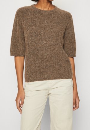 Personne portant un pull court en laine marron à manches courtes et un pantalon beige clair, debout devant un fond clair uni.