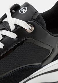 Črne superge, ki združujejo gladko usnje in teksturirano tkanino, bele vezalke in srebrno logotipno podrobnost na strani.