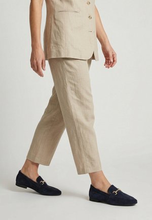 Person trägt beige verkürzte Hose und passenden Blazer, geht in marineblauen Wildlederloafern mit goldenen Schnallen vor einfarbigem Hintergrund.