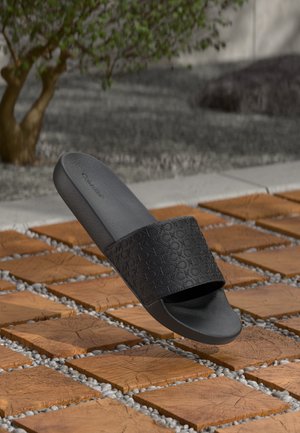 Czarne klapki z fakturowanym paskiem, z powtarzającym się wzorem, na drewnianych kafelkach z dodatkami żwiru. Gładka wkładka i minimalistyczny design.