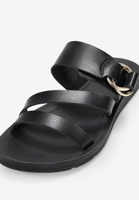 Sandalo in pelle nera con tre cinturini regolabili e un accento in metallo dorato circolare. Suola piatta e design minimalista.