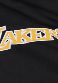Schwarzer Stoff mit gesticktem "LAKERS"-Text in kräftigem Gelb, umrandet in Weiß. Die Buchstaben haben eine strukturierte Optik.