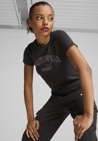 Melnā īsā piedurkņu t-kreklā ar košiem "SPORTSWEAR" uzrakstiem un logotipu priekšā, nēsāts kopā ar melnām biksēm. Gluda auduma tekstūra.