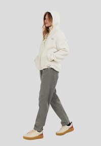 Kvinde går iført en hvid fleece hoodie, grå bukser og cremefarvede sneakers med gummisåler og røde hæl-accenter.