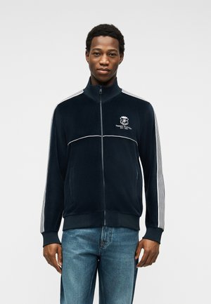 Bărbat purtând o jachetă sport neagră cu fermoar, dungi albe pe mâneci și logo pe piept, asortată cu blugi albaștri, stând în fața unui fundal alb.