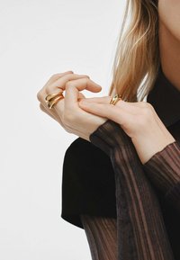 Anillos de oro de diferentes anchos en los dedos, con un acabado suave y acentos decorativos. Manga de una blusa con textura y rayas transparentes.