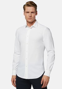 Chemise blanche à manches longues avec un col classique et un ourlet droit, dotée d'une fermeture à boutons sur le devant et d'une texture de tissu tissé lisse.
