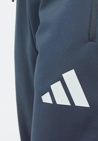 Námornícke jogger nohavice vyrobené z mäkkého materiálu, s výrazným bielym logom Adidas na stehne a so zipsovými vreckami po stranách.