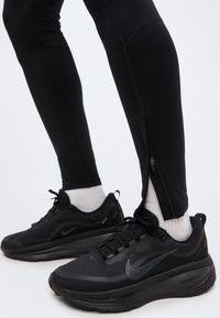 Πόδια που φορούν μαύρο αθλητικό κολάν με φερμουάρ στους αστραγάλους, άσπρες κάλτσες και μαύρα παπούτσια τρεξίματος Nike σε λευκό φόντο.