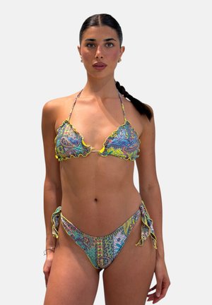 Donna con capelli scuri intrecciati che indossa un bikini colorato con motivo paisley e bordatura gialla, in piedi su sfondo chiaro e semplice.