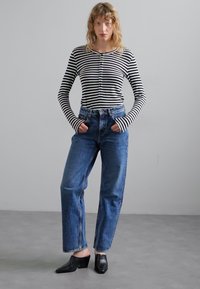 Randig långärmad topp tillsammans med högmidjade blå denimjeans. Svarta klackskor med blockhäl. Modell som står med händerna i fickorna, neutral bakgrund.