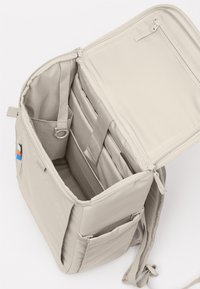 Sac à dos beige ouvert, montrant un compartiment rembourré pour ordinateur portable avec sangle de sécurité, poche intérieure zippée sur le couvercle et poches latérales à glissière.