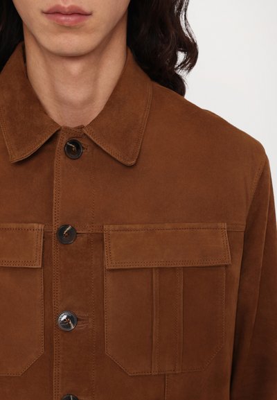 Brun ruskindsjakke med spidse kraver, fire frontlommer og sorte knapper. Har en glat tekstur og et enkelt, struktureret design.