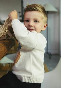 Sweter dzianinowy w kolorze kremowym z kolorowymi wzorami, w tym niebieskim kwiatem i brązowym akcentem. Czarne spodnie, siedzący przy drewnianym stole.