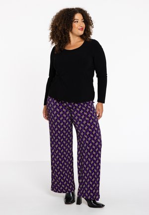Zwarte lange mouw top met een geknoopt detail bij de taille. Paarse wijde broek met een herhaald patroon van cirkels en accenten.