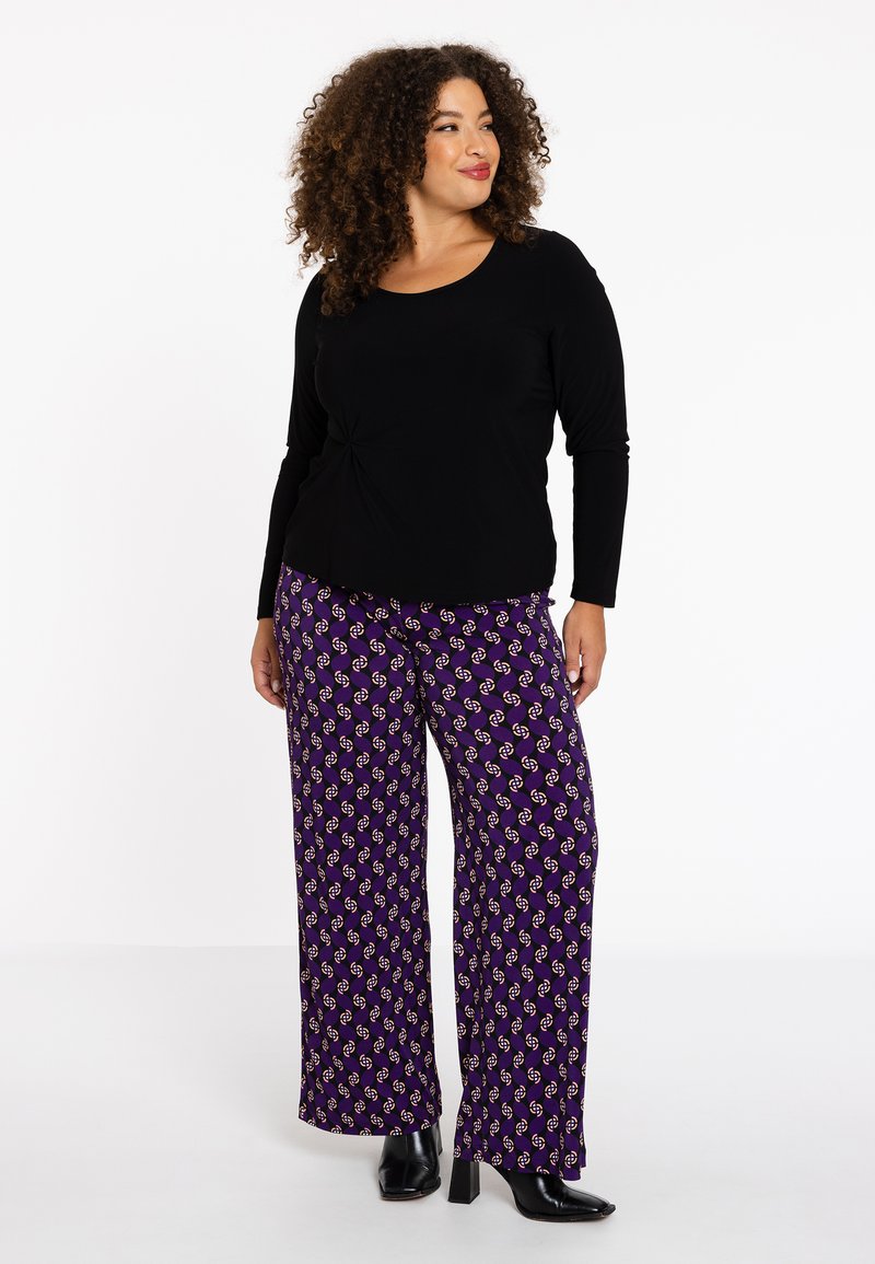 Zwarte lange mouw top met een geknoopt detail bij de taille. Paarse wijde broek met een herhaald patroon van cirkels en accenten.