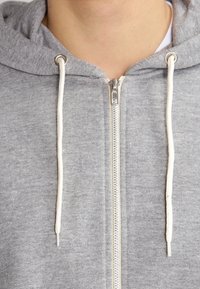 Primer plano de una persona con una sudadera gris con cremallera que tiene cordones blancos y un cuello y parte del cuello parcialmente visibles.